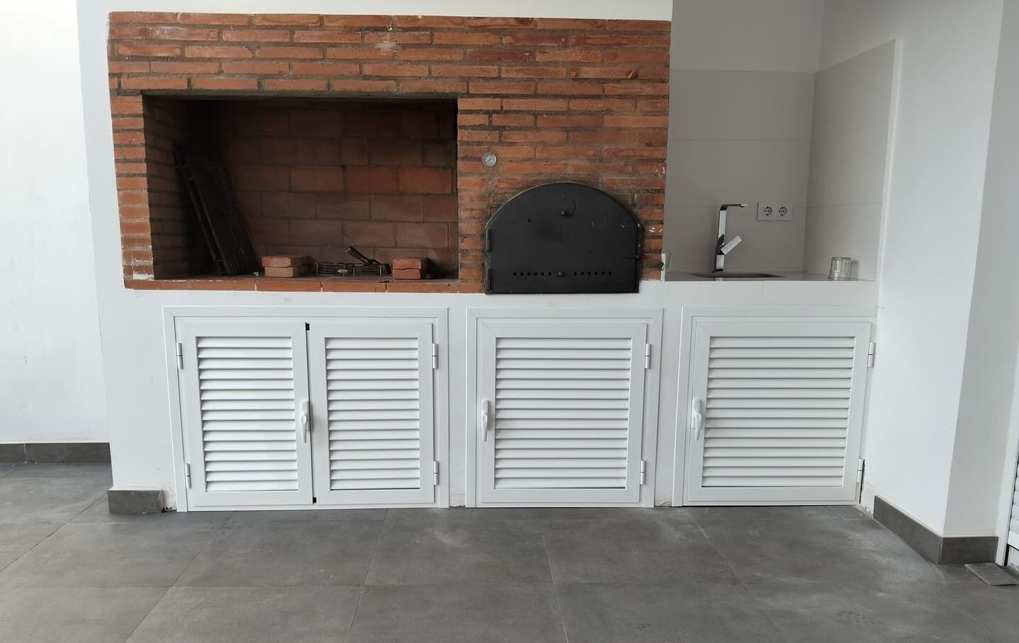 Puertas de aluminio blanco tipo lama bajo una barbacoa de obra, ajustadas al hueco